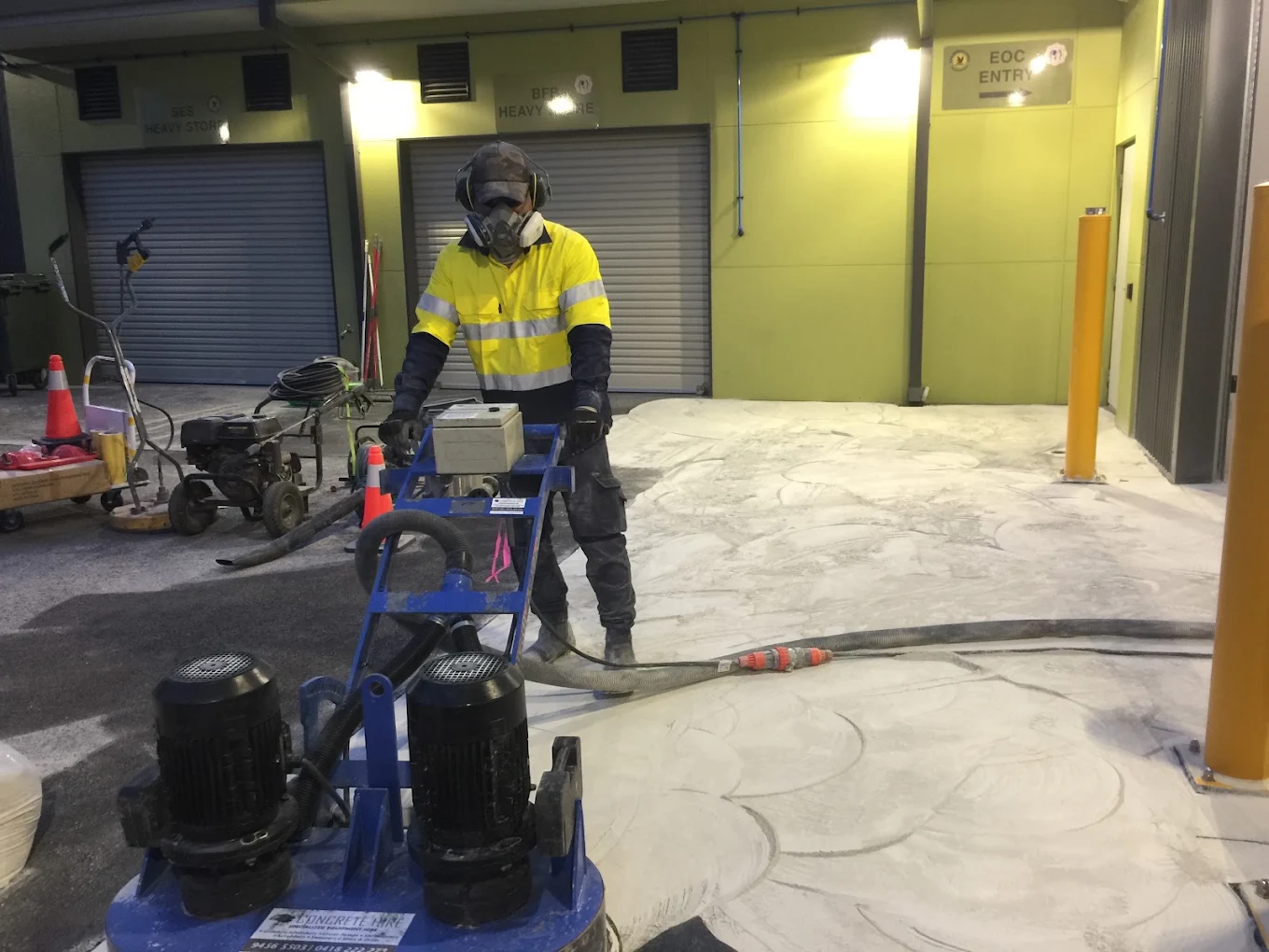 Operator in full PPE running a twin-head concrete grinder to prepare a commercial floor before coating