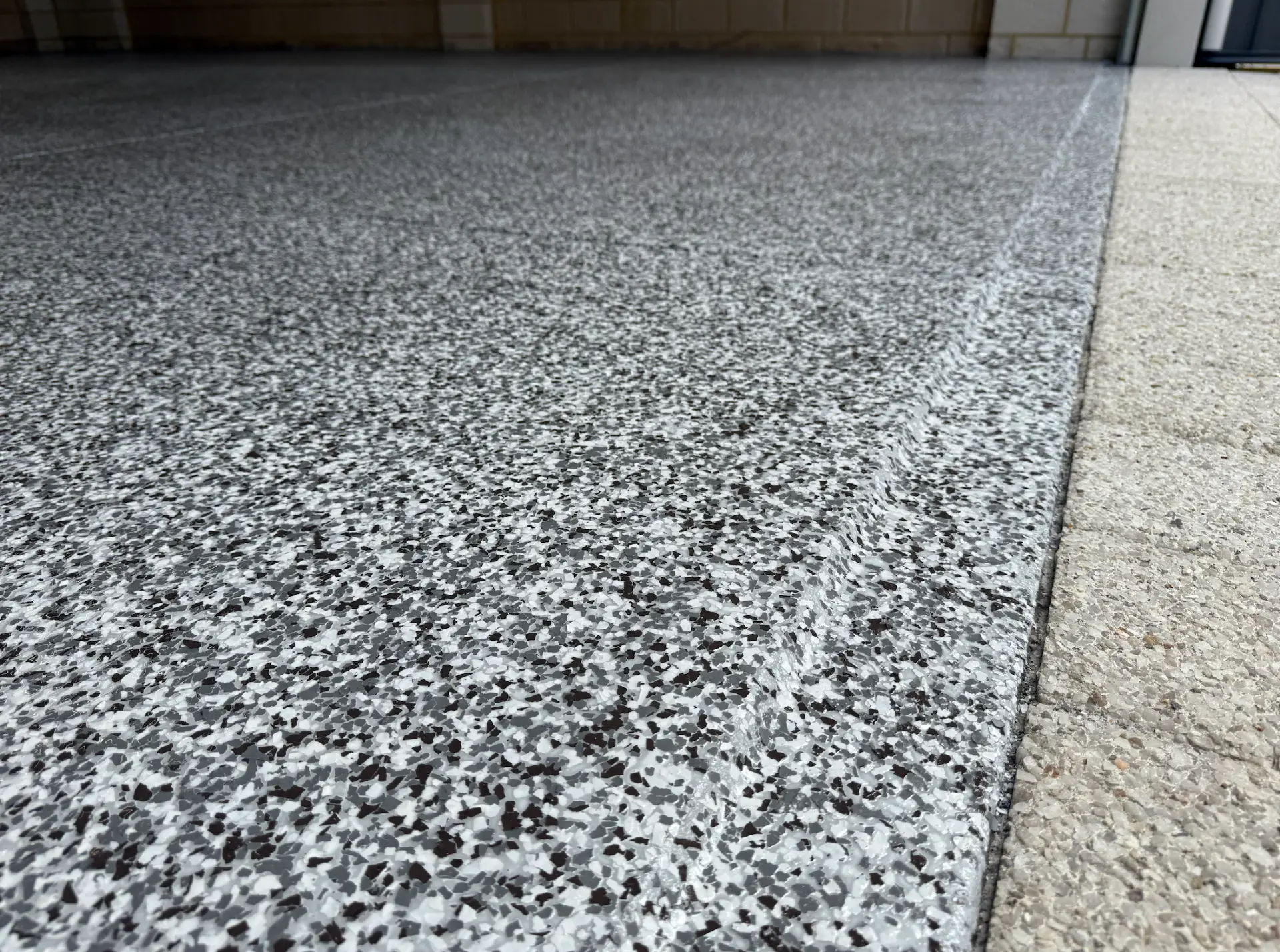 Grey and white epoxy flake floor coating with a speckled finish meeting exposed aggregate at a garage entry in Perth