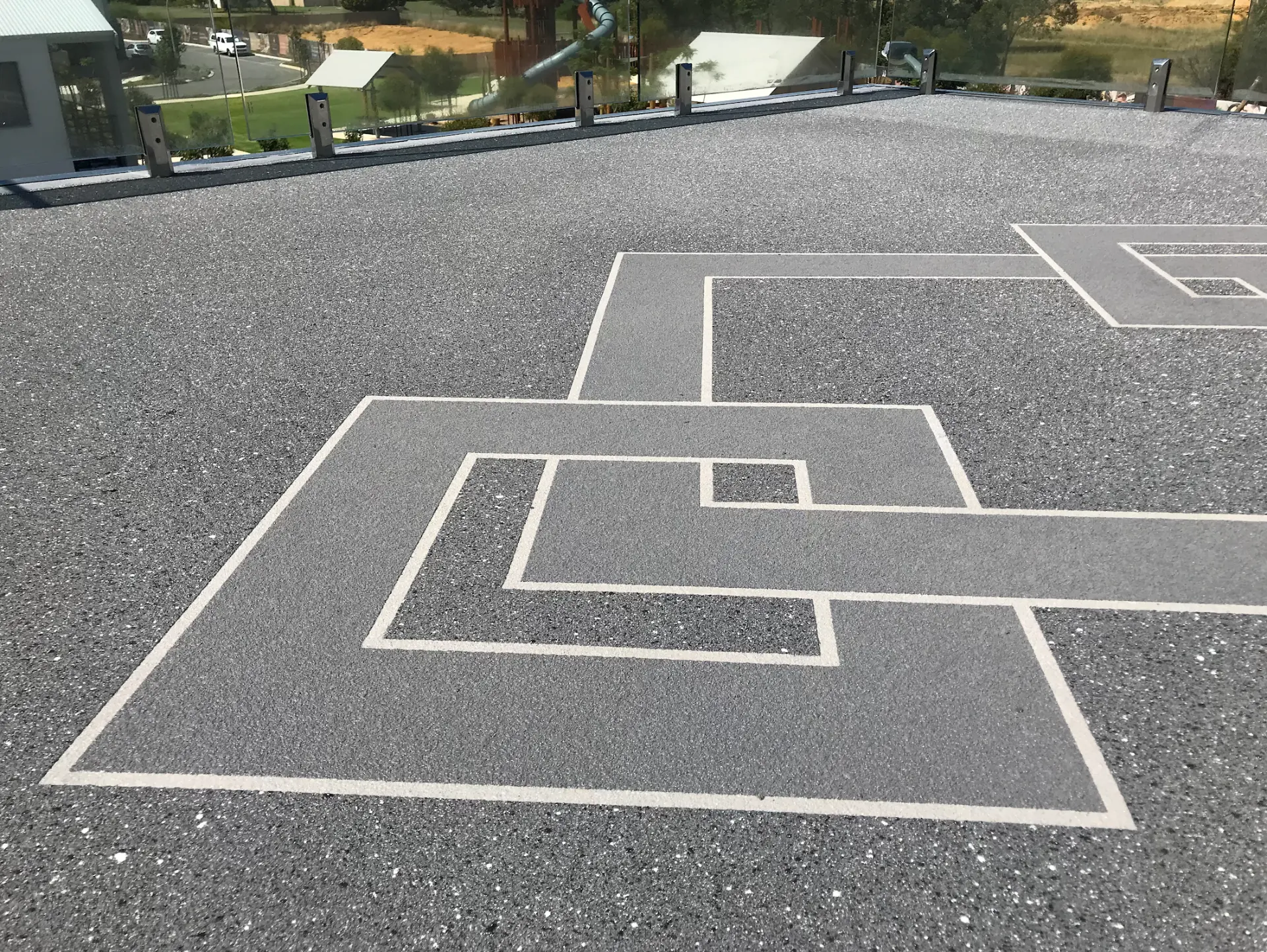 Grey resin-bound aggregate deck with a geometric maze pattern and pale border stripes behind a frameless glass balustrade at a Perth home